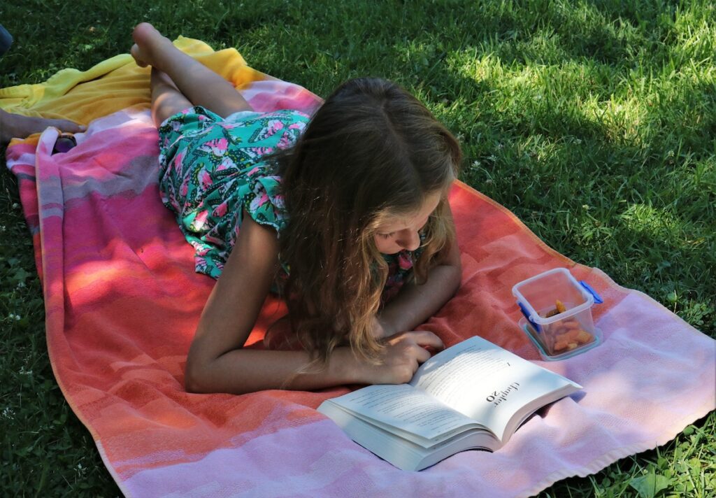 Young girl laying on a towel in the grass reading a novel